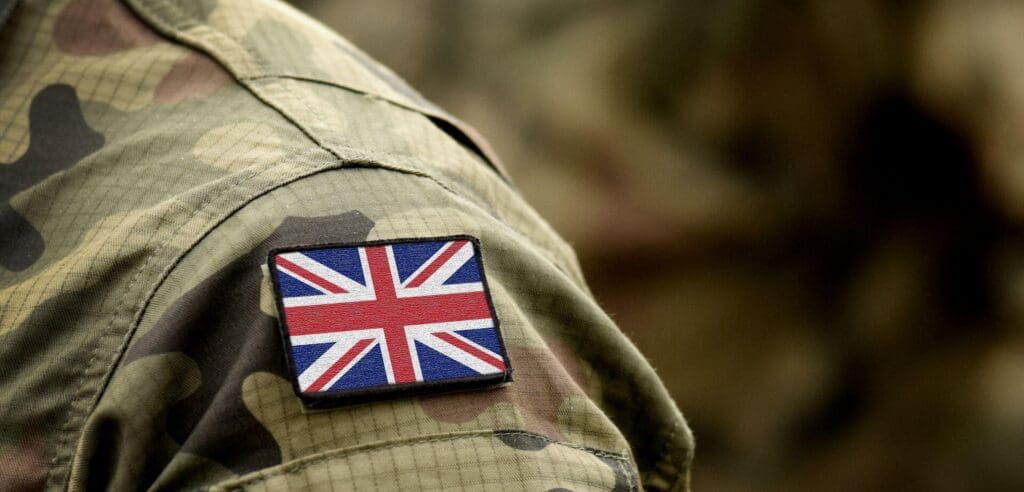 Close-up of a person wearing a camouflage military uniform with a Union Jack patch on the sleeve, spotlighting their dedication to UK military training. The patch is prominently displayed, symbolizing commitment. The background is blurred, emphasizing the focus on their attire.