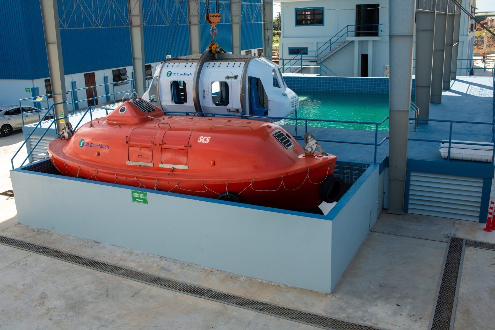 A scene at a training facility in Guyana features a red enclosed lifeboat and a helicopter simulator, positioned in front of an indoor swimming pool. Used for safety and emergency training exercises, both are situated on blue-tiled platforms.