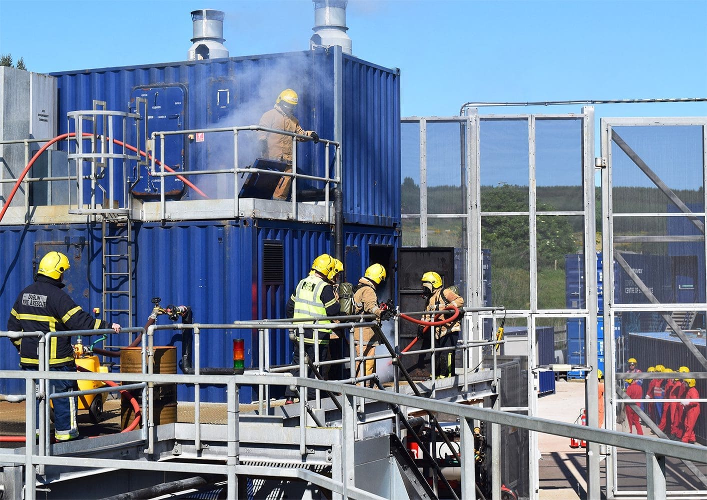 Firefighters in full gear are conducting a training exercise on a blue metal structure in Aberdeen. Some are using hoses to spray water, while others are climbing ladders. Smoke is visible at the top of the structure, and other firefighters are observing the scene.