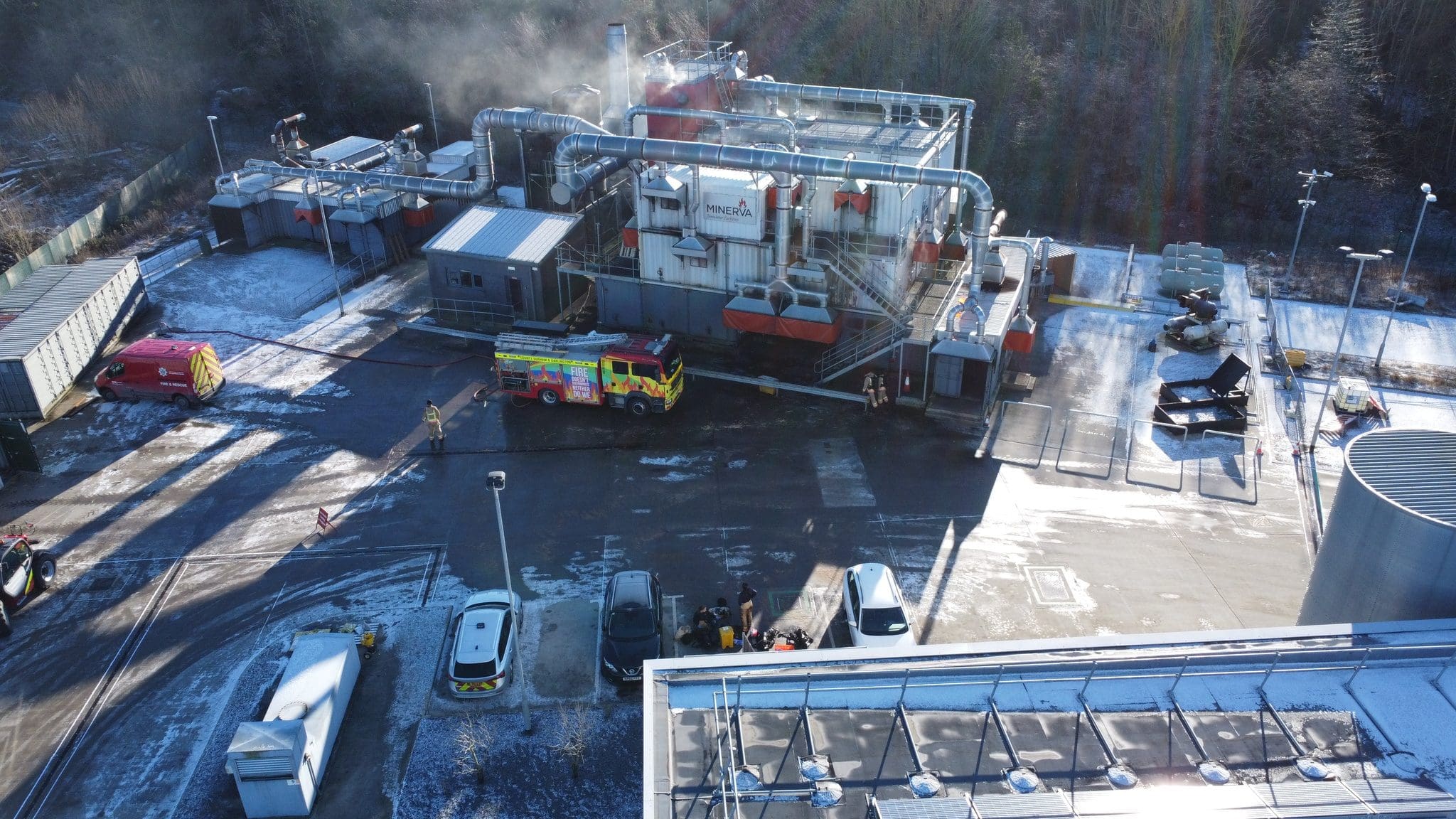 Aerial view of an industrial facility in Durham with a fire truck parked nearby, releasing some smoke. There are various structures, containers, and vehicles in the area, with some snow on the ground and surrounding trees.