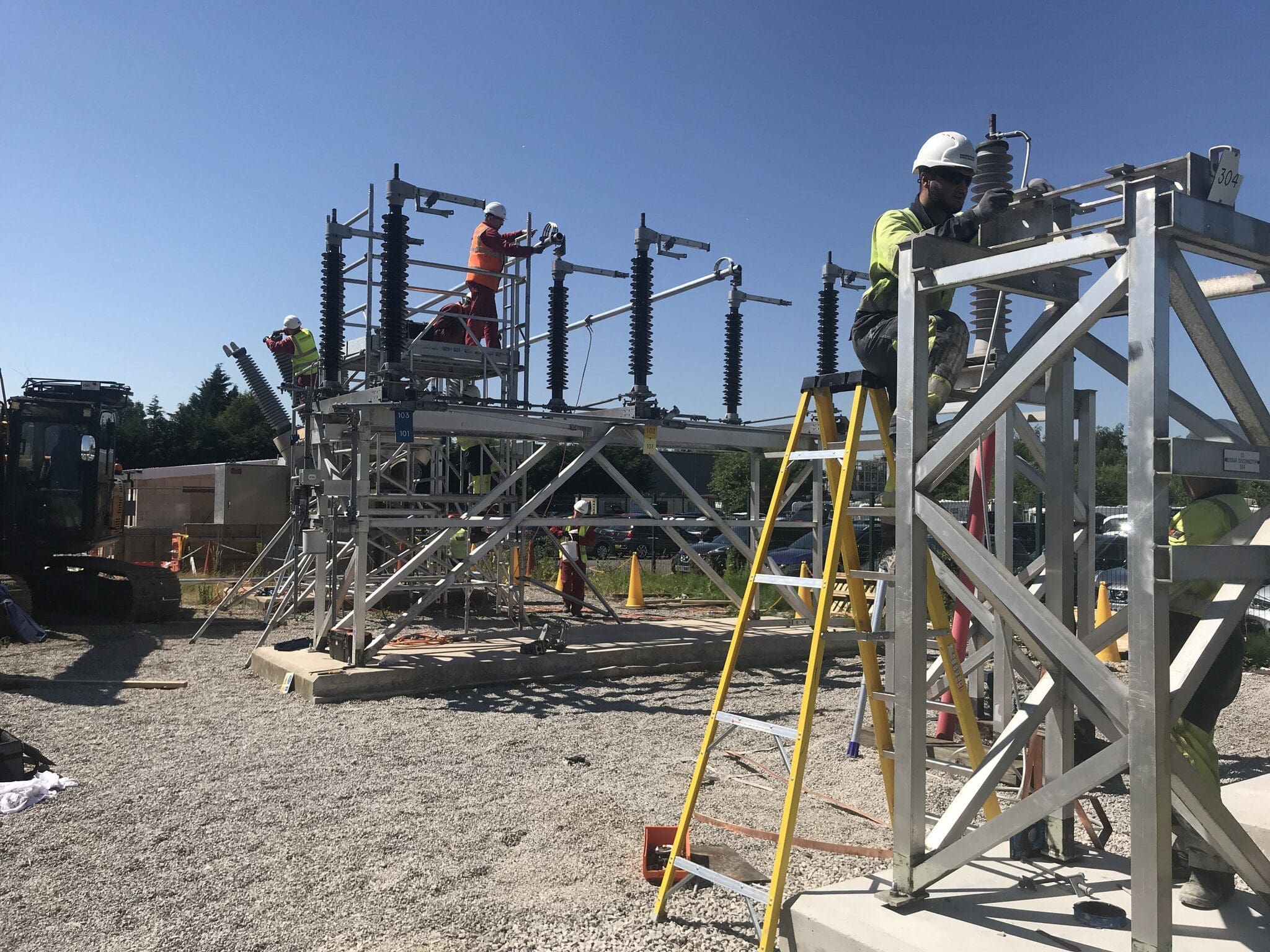 Construction workers are assembling a metal structure at a worksite in London. Several workers are seen on different levels of the structure while one stands nearby. Equipment, including ladders and machinery, surrounds the area, with a clear blue sky in the background.