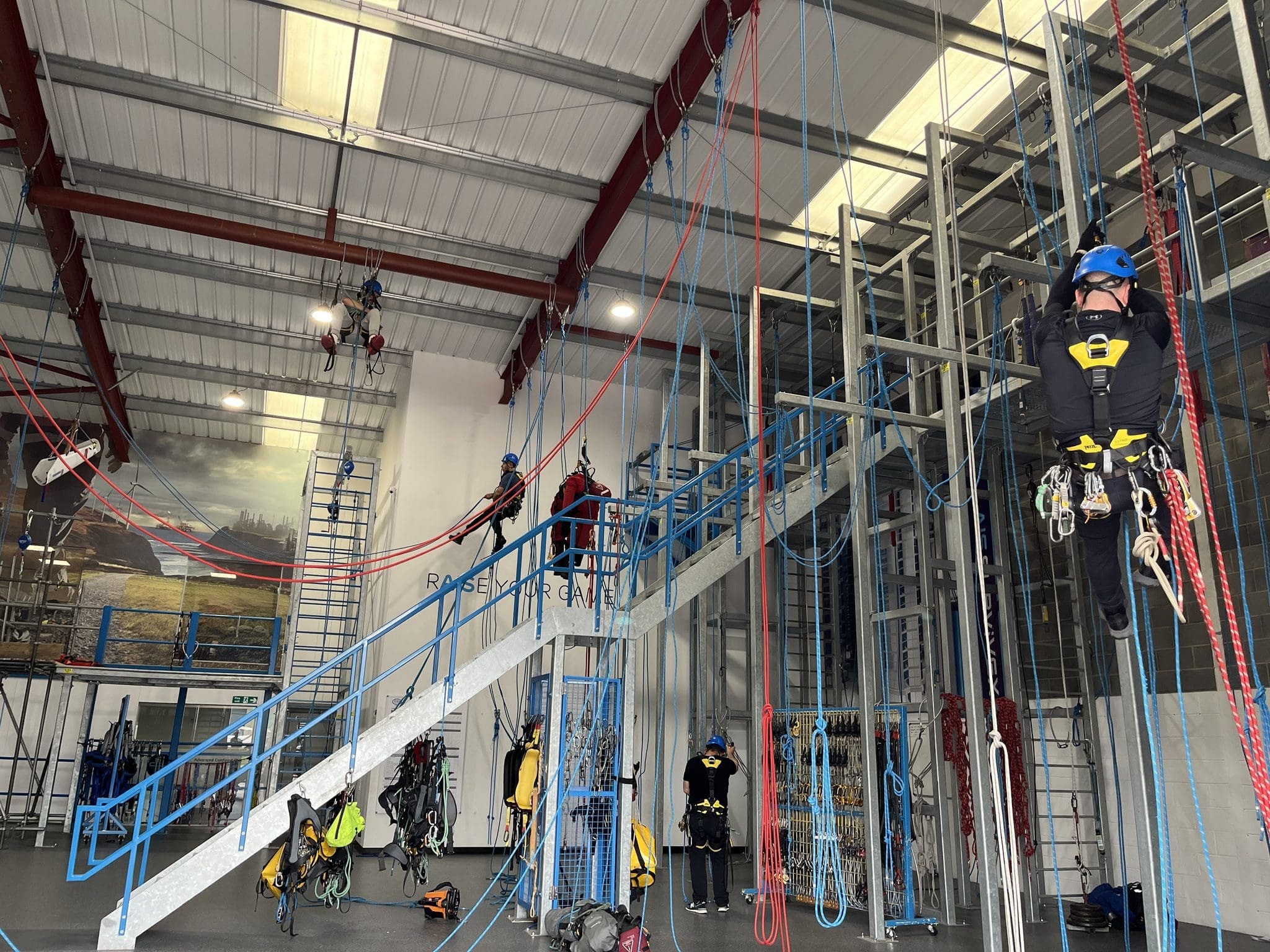 Indoor training facility in Newcastle with multiple individuals practicing rope access techniques. One person is ascending a rope while others are either climbing or observing. Various ropes and climbing equipment are set up throughout the space, which includes a staircase and metal structures.