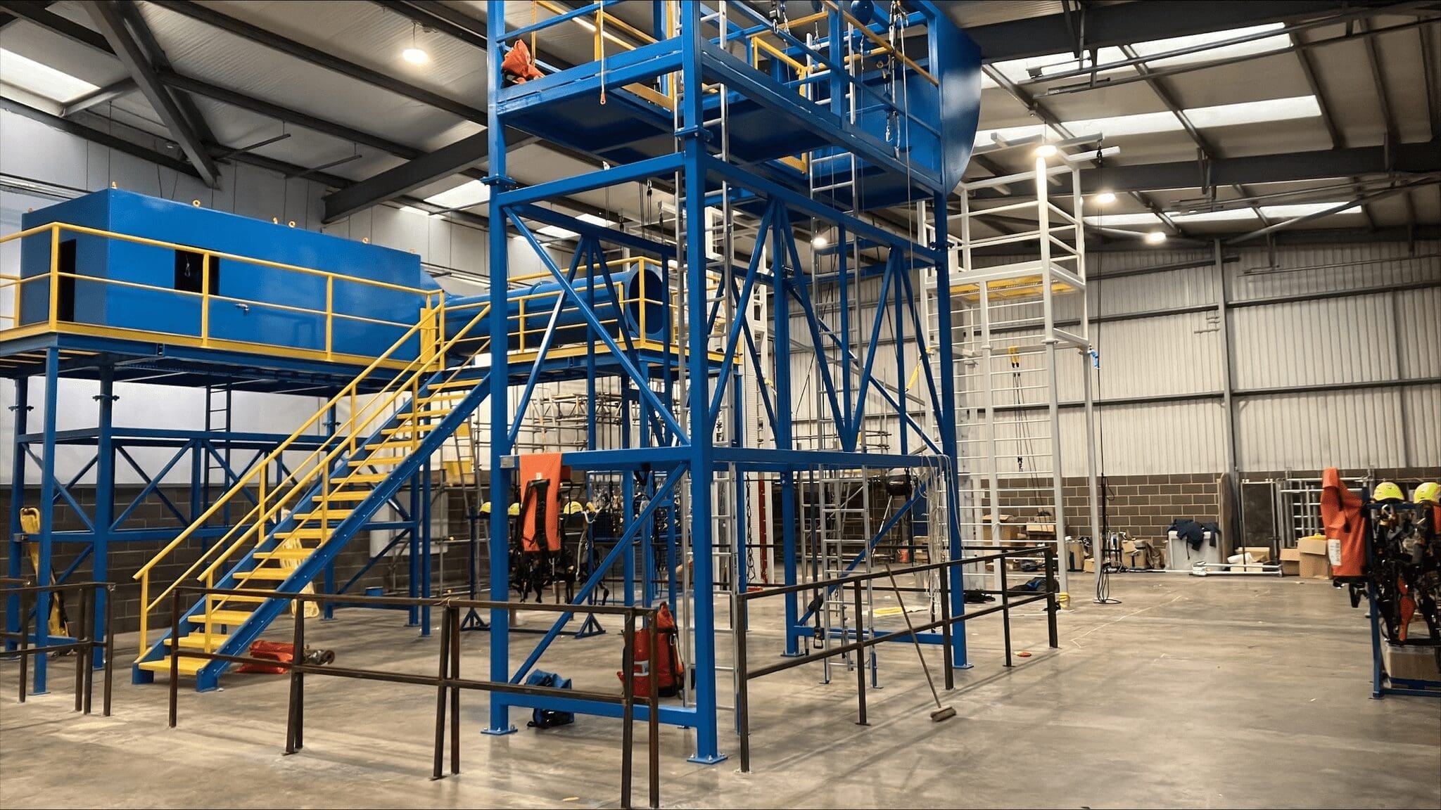 A large industrial warehouse interior in Teesside with blue metal structures and yellow stairs. The space includes various equipment and machinery, metal railings, and workers in safety gear. The ceiling has metal beams and skylights, and there are storage areas along the walls.