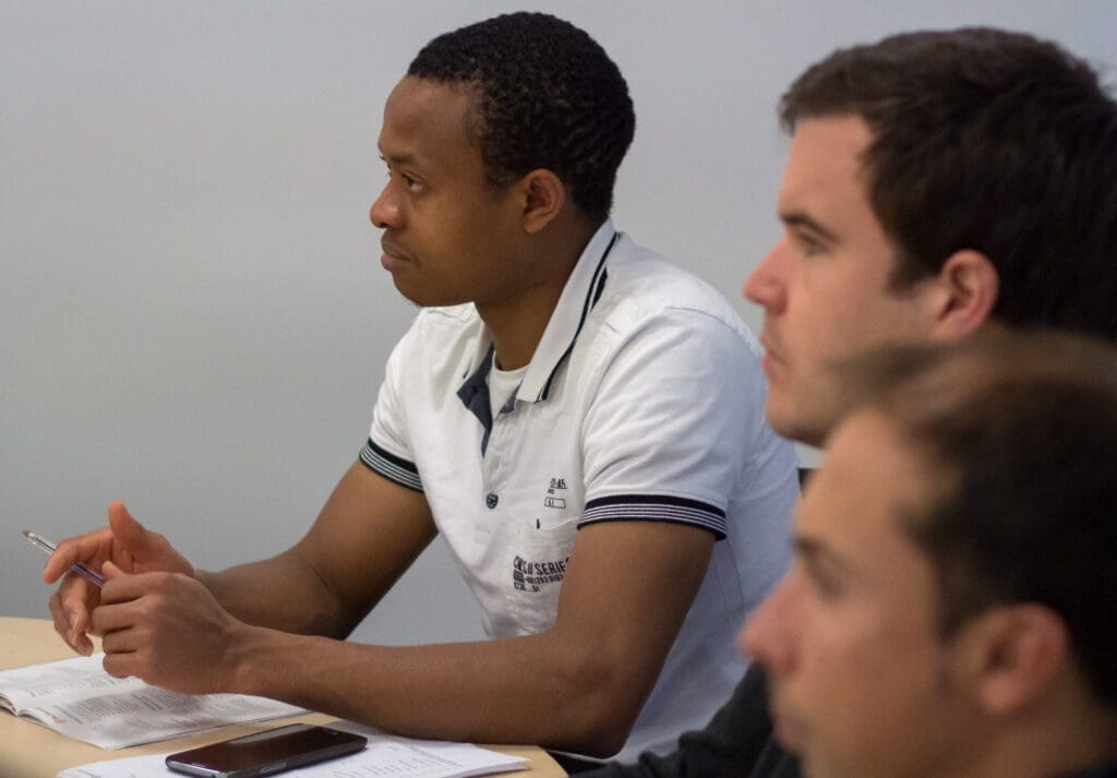 Three people are seated in a row at a table, appearing to be attentively listening. The person closest is slightly blurred, while the two individuals farther are more in focus. Open books and a pen suggest a study or meeting environment, reflecting our services in fostering collaborative learning.