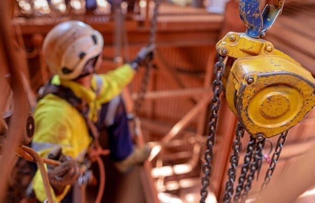 A construction worker, likely an offshore rigger new to the job, wearing a safety helmet and high-visibility clothing sits and works amidst scaffolding. The foreground shows a yellow industrial chain hoist with a metal hook. The background appears to be an industrial or construction site.