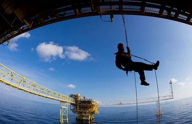 A person in safety gear is suspended by a rope under a large metal structure, possibly an offshore oil rig. Utilizing Offshore Rope Access techniques, they navigate the rig against the backdrop of a blue sky and ocean, with several yellow platforms extending over the water.