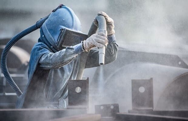 A person wearing a protective suit and helmet operates a sandblasting tool, creating a cloud of dust. Various metal structures are in the background, suggesting an offshore maintenance operation. The scene appears to be an industrial or construction setting.