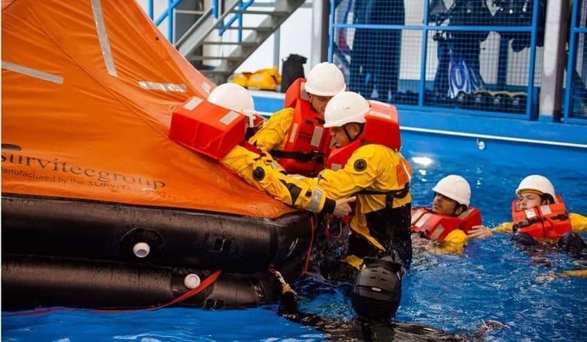 A group of mariners in yellow suits, life vests, and helmets participate in a water safety training exercise by 3t Training Services. They are in a pool, securing themselves to an orange inflatable life raft. A metal rail and stairs are visible in the background.