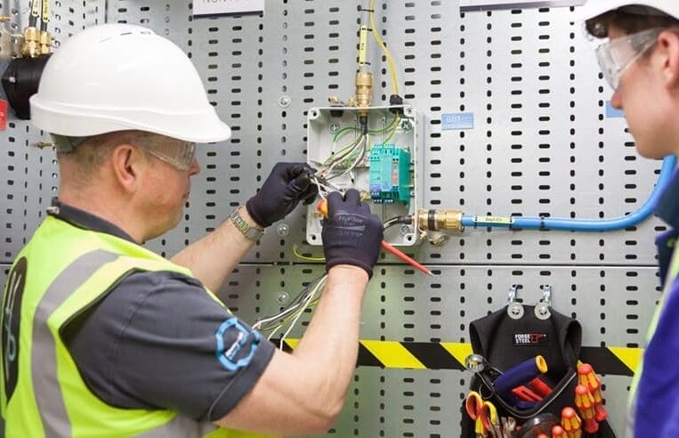 Two individuals wearing safety helmets and reflective vests work on wiring inside a metal panel, showcasing their CompEx competencies. One uses tools to manipulate wires, while the other observes. Various tools and cables are attached to the wall beside them, reflecting their rigorous 3t Training Service standards.