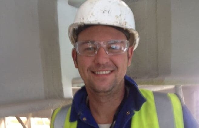 A man named Lee, wearing a white hard hat, safety glasses, and a high-visibility vest, smiles at the camera. Dressed in a blue shirt, he appears to be in an industrial or construction setting. Having transitioned from his former bricklayer job, Lee is now thriving in his offshore career.