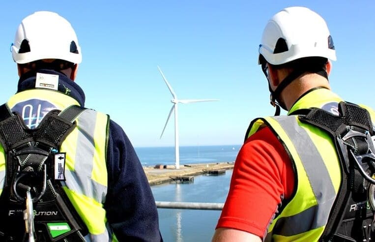 Two workers in high-visibility vests and helmets are observing a wind turbine near a body of water. One worker, registered with WINDA, is wearing a navy blue top, the other in red. Both are equipped with safety harnesses as they follow GWO protocols under a bright, clear sky.