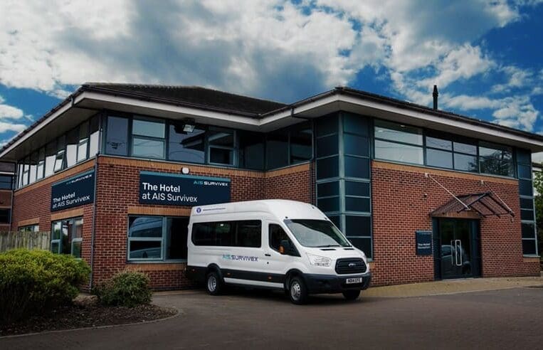 A brick building with large windows houses "The Hotel at AIS Survivex," known for its exceptional training services. A white van with "AIS Survivex" branding is parked in front. The setting is under a partly cloudy sky, and there are some bushes beside the building.