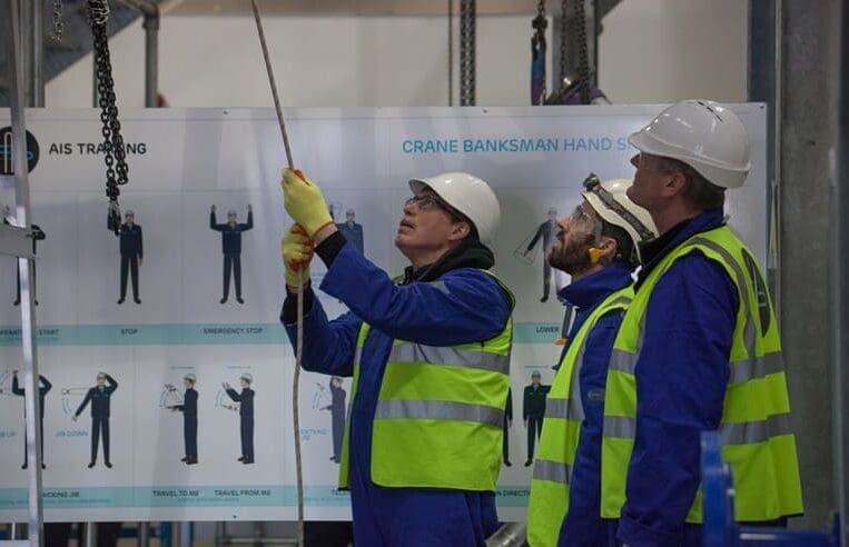 Three workers with OPITO Rigging Accreditation, wearing high-visibility vests and hard hats, are operating a crane inside an Aberdeen work facility. One worker pulls on a rope while the others observe. Behind them is a large sign displaying crane banksman hand signals from 3t Training Services.