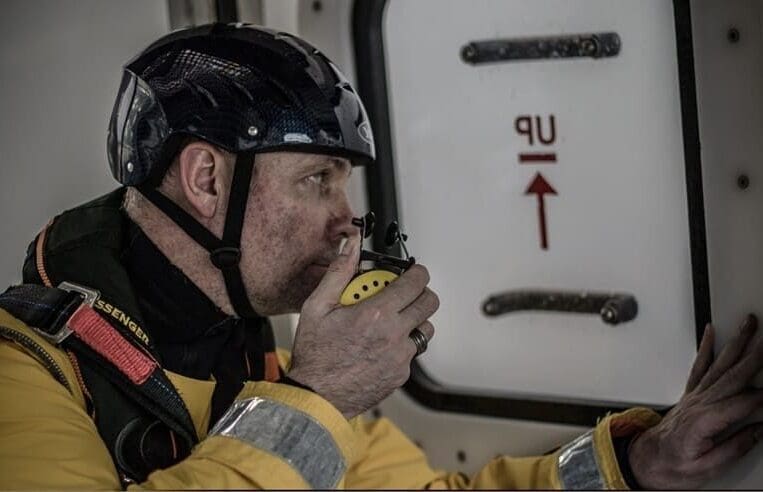 A person wearing a helmet and a yellow protective suit holds a radio to their mouth while looking out a window inside a vehicle or structure. A sign on the window reads "UP" with an arrow pointing upward, suggesting training in HUET.