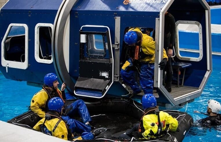 A group of people in blue helmets and yellow jackets, trained by 3t Training Services, are participating in a water survival training exercise. They are exiting a mock spacecraft module on a pool and climbing onto a floating black raft. One person assists from the raft while another stands in the water.