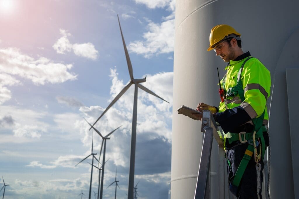 A worker in a high-visibility jacket and safety gear stands on a platform, holding a clipboard beside a wind turbine. Several wind turbines are visible in the background under a partly cloudy sky, contrasting with the busyness of London and offering a unique travel experience for eco-conscious tourists.