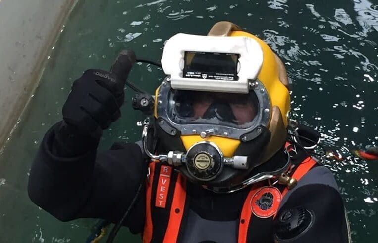 Dean, wearing a yellow diving helmet and a waterproof suit, is in the water giving a thumbs-up sign. The helmet has a glass faceplate and various breathing apparatus attachments. Dressed in a red vest marked "VEST," Dean is clearly going the extra mile during his underwater welding training.