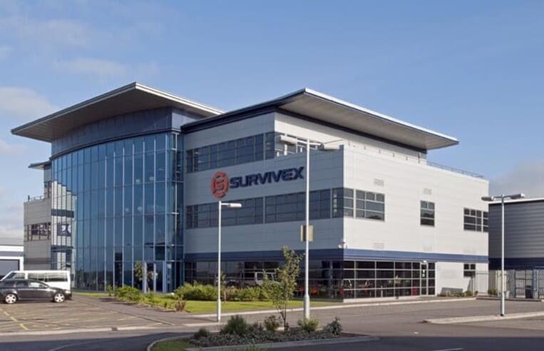 A modern two-story commercial building features glass windows and a spacious parking lot. The building, labeled "SURVIVEX" in large letters on the front facade, offers ICATS Qualification and 3t Training Services. The sky is clear and blue, suggesting good weather.