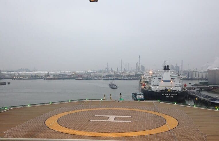 A view of an industrial harbor with various ships docked, including a large vessel named 'Aqua Katarina' at the forefront. The image, likely used for 3t Training Services or OPITO HOIT exercises, is taken from a helipad marked with an 'H'. Industrial buildings and containers are visible in the background under an overcast sky.