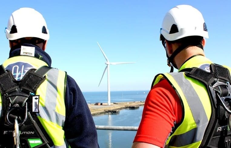 Two workers wearing safety helmets, high-visibility vests, and safety harnesses stand on an elevated platform overlooking an offshore wind turbine. The background features a clear blue sky and the ocean—a scene exemplifying the growing global wind sector.