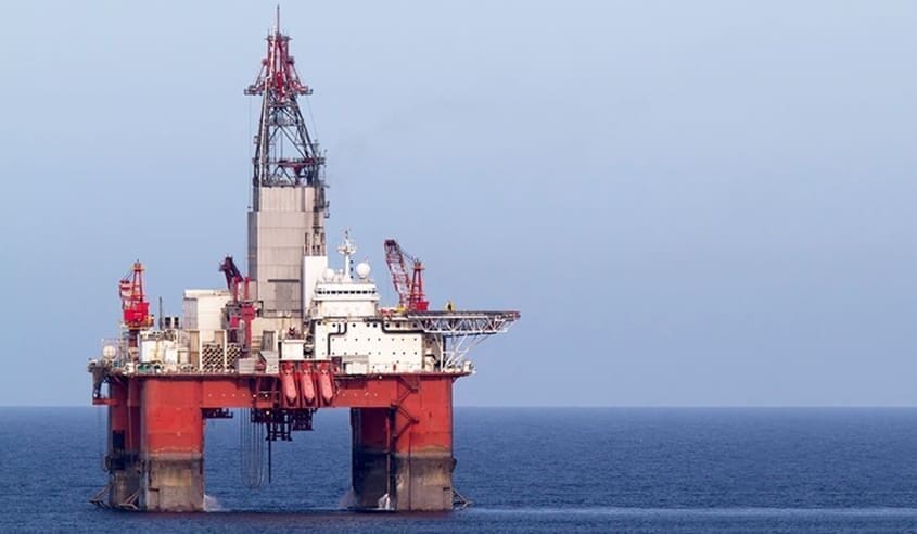 An offshore oil rig is positioned in the ocean against a clear sky. The platform, crucial to the oil and gas industry, features red and white structural components with cranes and other equipment visible on its deck. Guided by large legs that extend into the water, it stands tall and operational.