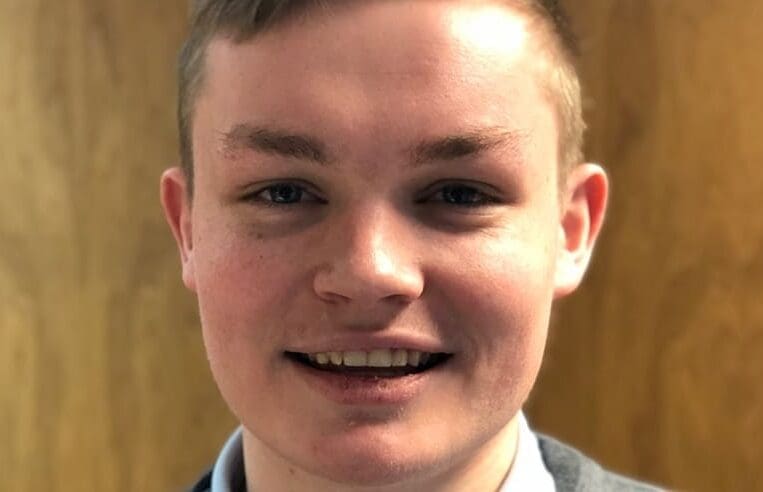 A close-up photo of a young person with short, light brown hair. They are smiling softly and wearing a collared shirt, perhaps contemplating their career plans. The background is blurred but appears to be a wooden surface.