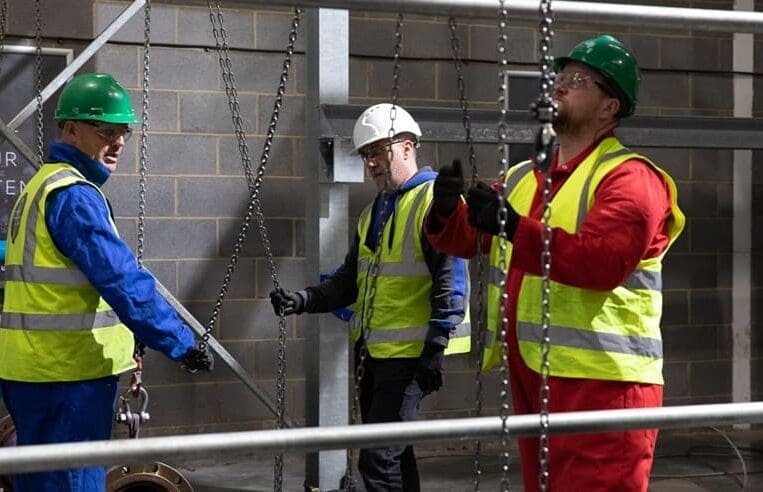 Three construction workers in safety gear, including helmets and high-visibility vests, operate chains in an industrial setting. The worker on the right is in red overalls, while the other two are in blue. Focused on their tasks, these professionals exemplify what it means to lift your career to new levels as riggers.