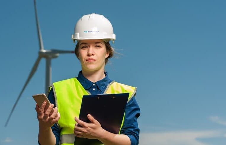 A person wearing a white hard hat and a yellow reflective vest stands outdoors in front of a wind turbine, symbolizing the transition to green energy. They hold a tablet and a clipboard, looking forward under the clear blue sky.