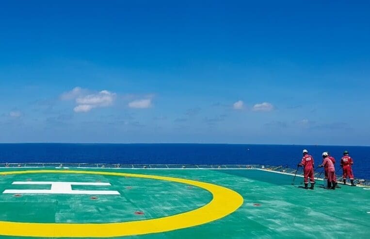 Two workers in red coveralls and helmets stand on a helipad with a yellow circle and white "H" marking. The helipad is on a large vessel or offshore platform, with the ocean and blue sky in the background, signaling the Kick Off for Shutdown Season 2022.