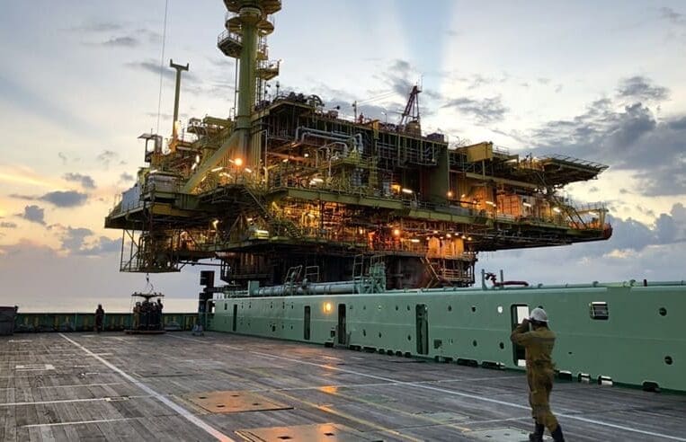 A worker in protective gear walks on the deck of an offshore oil platform at dusk. The platform is illuminated with numerous lights and surrounded by a cloudy sky and calm sea. For those starting their first offshore job, it's essential to familiarize yourself with the complex structure, levels, pipes, and equipment.