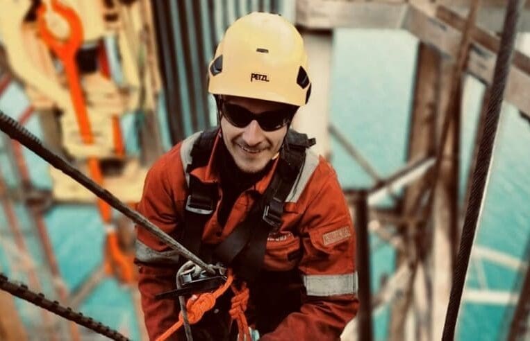 Daniel, a military veteran in a yellow helmet, sunglasses, and red protective gear, is performing work at height offshore. Using ropes and safety harnesses while positioned on a metal structure with a body of water in the background, he truly embodies his dream job.