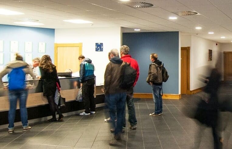 People are standing and interacting at a reception desk in a modern, brightly-lit office or public space with dark tiled floors and blue accent walls. Some are being attended to by staff members, discussing job opportunities and growth through 3T Training Services, while others wait in line or walk by.