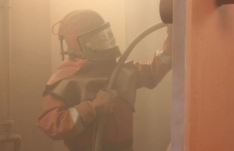 A person wearing protective gear, including a helmet and mask, is using a hose to spray or clean a surface in a dusty or smoky environment. The area is filled with haze, suggesting cleaning or maintenance work in a potentially hazardous location, akin to the conditions faced in offshore jobs.