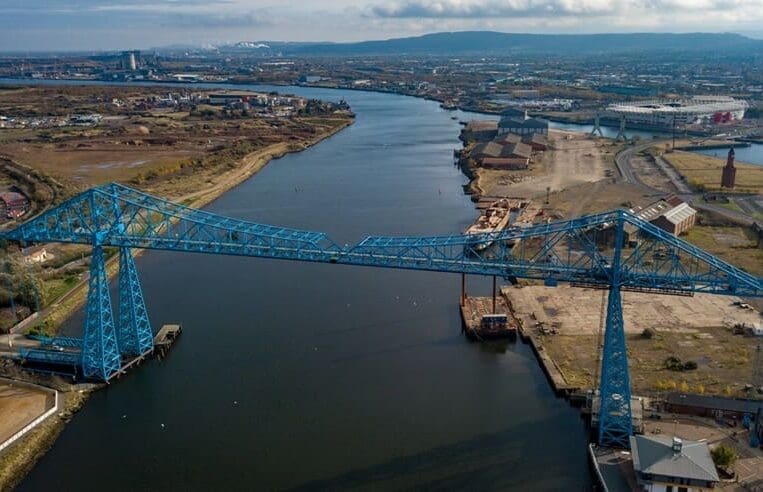 Aerial view of the large blue steel transporter bridge spanning a river in Tees Valley. The landscape includes industrial buildings and open land on either side, with a cloudy sky and hills in the background. The bridge's tall towers and cables are prominent in the image.