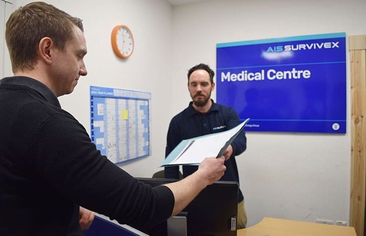 Two men are in a medical center. One man with short hair is handing a document to another bearded man behind the counter. A sign on the wall reads "AIS Survex Medical Centre," where HGV drivers often come for medical appointments. In the background, a clock and schedule chart are visible.