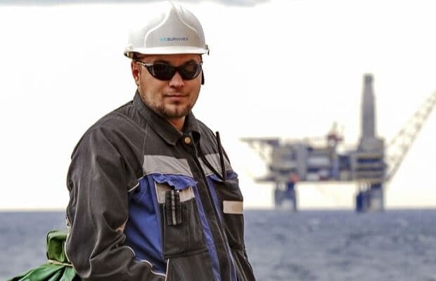 A person wearing dark protective workwear, a white safety helmet, and sunglasses stands near an offshore oil rig in the North Sea. The individual has a communication device clipped to their jacket and displays a neutral expression. The offshore platform is visible in the background across the water.