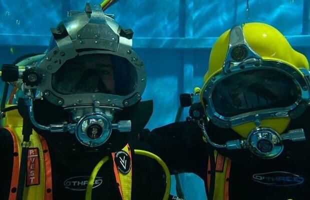 Two divers, possibly training for an offshore career, are underwater wearing full-face helmets and wetsuits. They pose side by side in what looks like a controlled environment or training pool. The blue pool wall forms the background, and both divers have visible breathing apparatus.