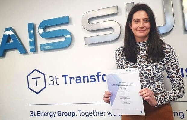 A woman with long dark hair is holding a certificate and standing in front of a wall featuring the logos and names "AIS Survivex" and "3t Energy Group. Together We." She is wearing a patterned long-sleeve top, smiling proudly, having just completed her OPITO-approved MIST training.