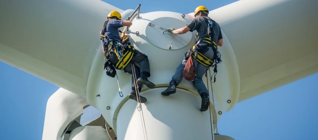 Two skilled technicians wearing safety gear are performing maintenance on a large wind turbine. They are secured with harnesses and ropes, and one is reaching towards the center hub while the other holds onto the structure. Their expertise ensures safety under a clear blue sky.