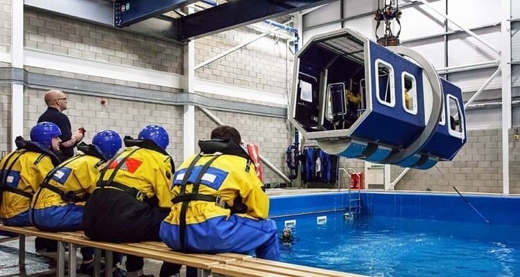 A group of survival experts in yellow and blue protective gear watches as a blue and white cabin module is lowered into an indoor pool by a crane, likely as part of offshore survival refresher training. The area is within a large industrial facility.