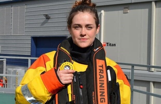 A woman in a yellow and orange high-visibility rescue suit stands in front of a grey building. She is holding a piece of equipment related to the In-water CA-EBS and wearing a life vest labeled "TRAINING." Her hair is tied up in a bun, and she has a neutral expression, prepared for her Fit to Train Assessment.