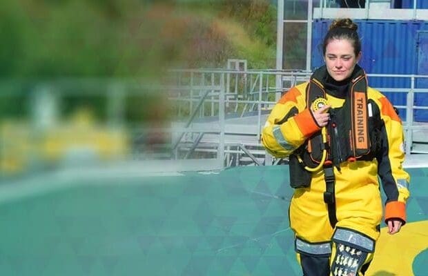 A person wearing a yellow and black waterproof suit with "TRAINING" written on the life vest walks on a platform, likely familiar with BOSIET requirements. The background includes a blue shipping container and industrial structures.