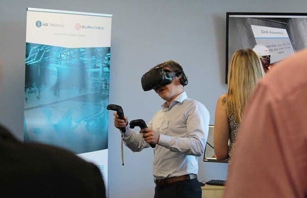 A man wearing a virtual reality headset holds VR controllers in both hands. He is in a room with other people, standing near a promotional banner for 3t Energy Group and a computer screen. Another person in the foreground is facing away from the camera.