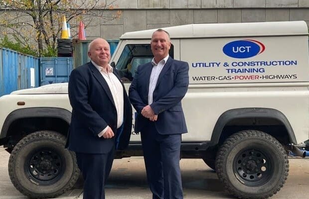 Two men in suits stand in front of a white utility vehicle with the logo and text "UCT, Utility & Construction Training, Water-Gas-Power-Highways" on its side. They are outside, with a blue container and safety cones visible in the background, representing 3t Energy Group's dedication to industry safety.