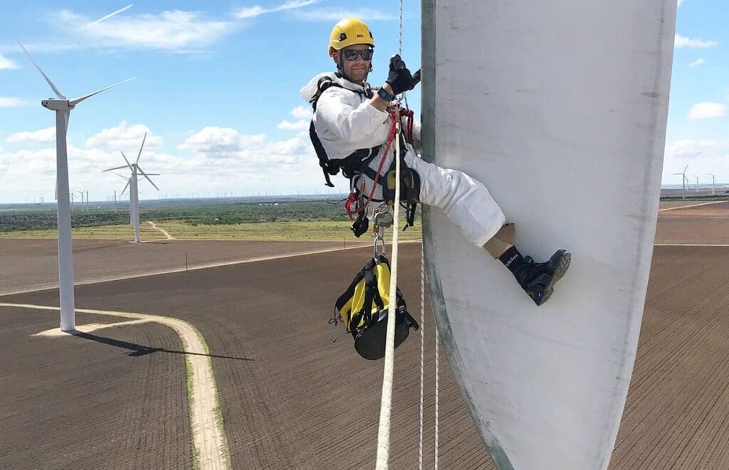 A worker in protective gear and a yellow helmet is suspended by ropes while performing maintenance on a large wind turbine blade. Several other wind turbines are seen in the background against a blue sky with scattered clouds, showcasing opportunities for those undergoing military transition to earn ELCAS Credits.