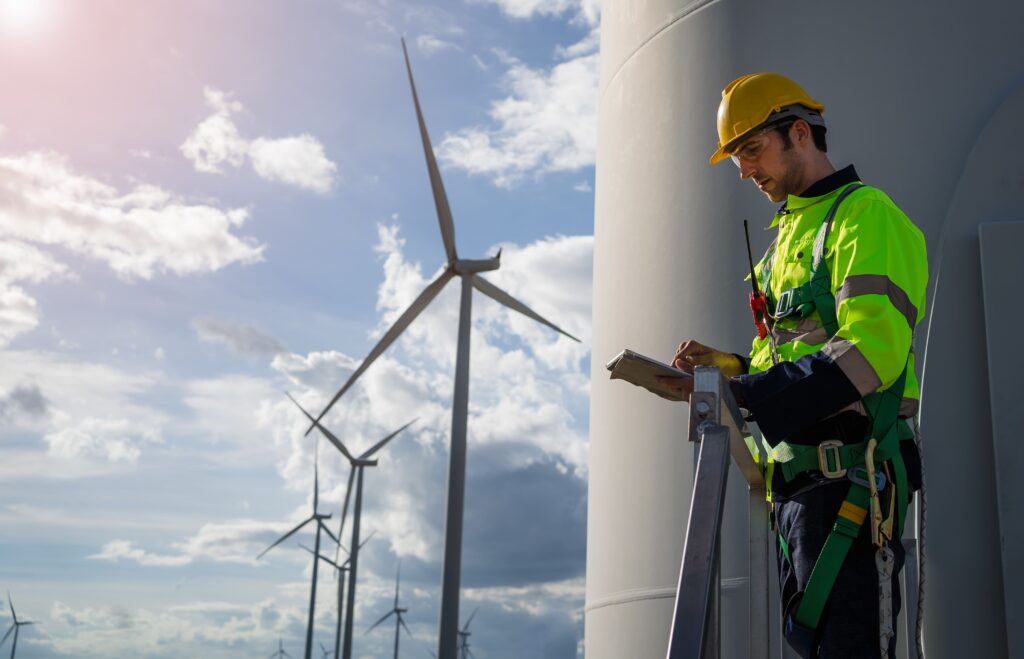 A person wearing a high-visibility jacket, helmet, and safety harness works on a tall structure with a tablet. Against a backdrop of wind turbines and a partly cloudy sky, this scene exemplifies the demanding tasks showcased at the wind industry careers event.