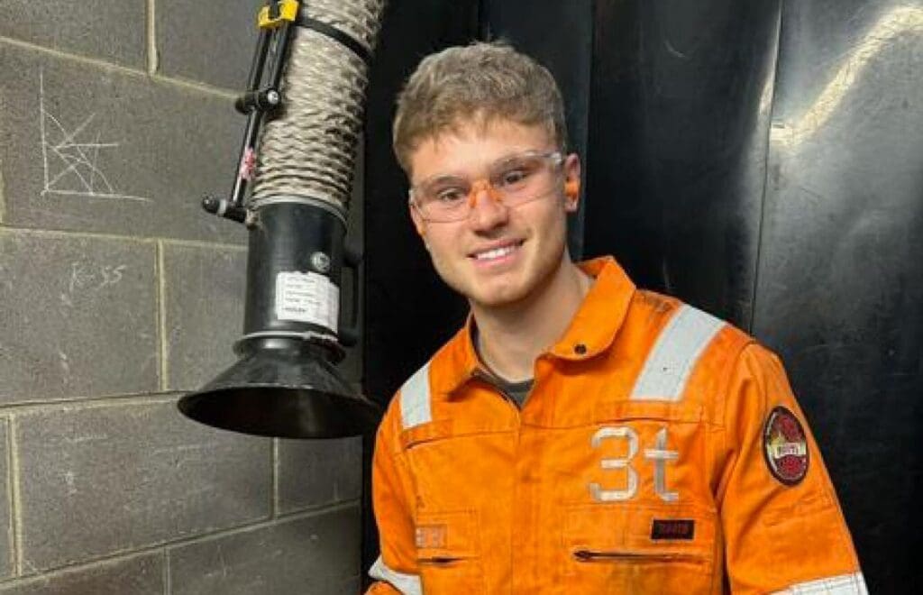 A man in an orange work jumpsuit labeled with "3t" stands in a workshop. He is wearing safety glasses and smiling, possibly proud after a prestigious competition. The background includes a brick wall and industrial ventilation equipment.