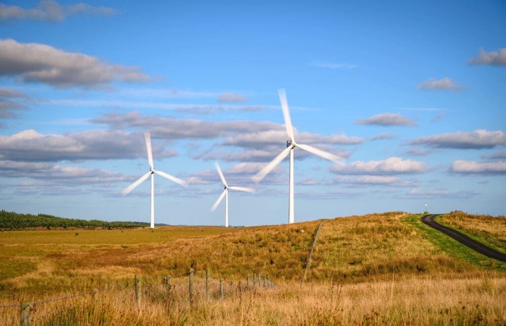 Three white wind turbines, a testament to the wind industry, stand in a row in a grassy field under a blue sky with scattered clouds. The blades are rotating, while a paved road runs through the field on the right side of the image.