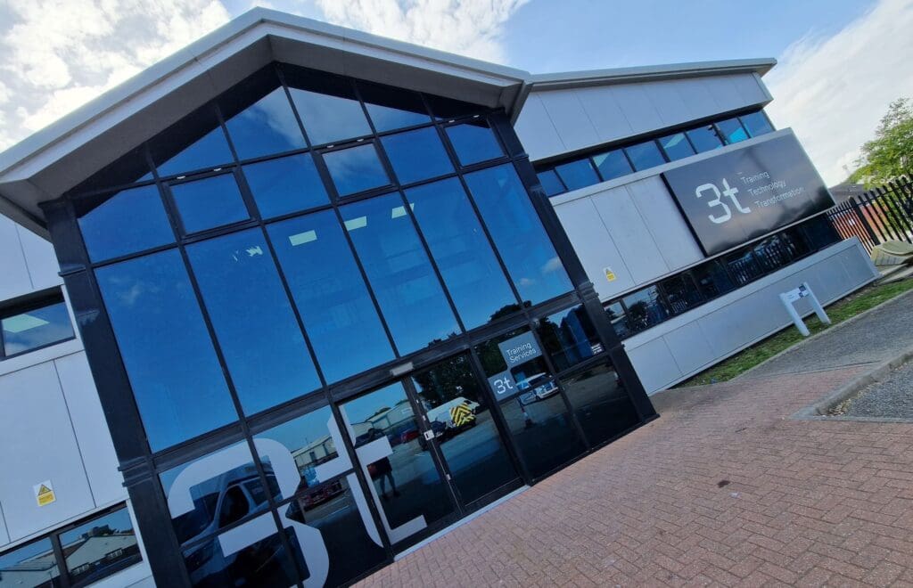 The image shows the exterior of a modern building with large glass windows. The building, part of the Middlesbrough training site, has the logo and text "3t Training Technology Transform" on a dark panel. The entrance features automatic glass doors, and there is a paved path along the front.