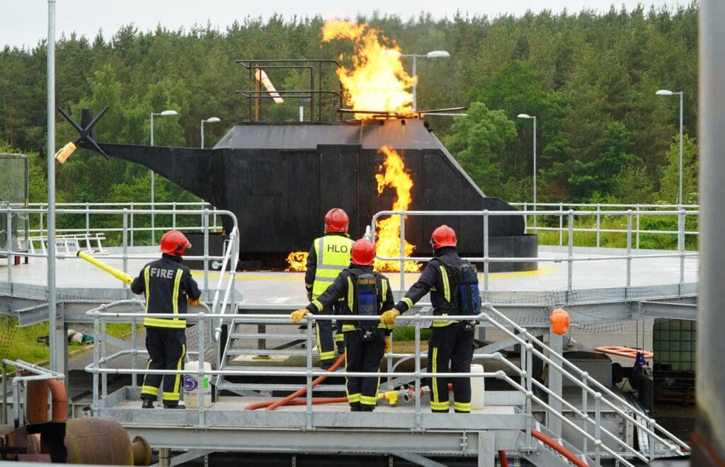 Four firefighters in protective gear stand on a metal platform, extinguishing a simulated helicopter fire with a hose. Flames and smoke rise from the helicopter structure at the Co Durham centre during helideck firefighting training. Trees are visible in the background.